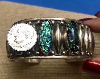 May include: Silver bracelet with three oval-shaped green and blue glass inlays. The bracelet has a silver coin with the words "Liberty" and "In God We Trust" and the year "2018" on it.
