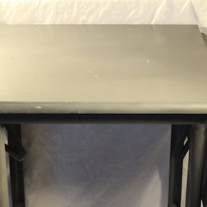 Pode incluir: Uma mesa dobrável cinza com uma superfície lisa e plana. A mesa é feita de madeira e tem um design simples e moderno.