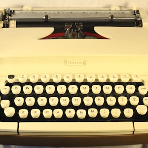 May include: Cream-coloured vintage typewriter with a full QWERTY keyboard and white keys. The typewriter has a red ribbon indicator and a brown base. The brand name is visible on the front.
