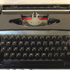 May include: A black Sears typewriter with a QWERTY keyboard layout. The machine features a glossy black metal body, a silver-toned front panel with the text Sears, and black keys with white letters. The internal typebars and red ink ribbon are visible.