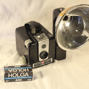 May include: A black Brownie Hawkeye camera with a flash attachment and a roll of Holga 400 black and white film. The camera has a silver lens and a black handle. The film box is black and blue with white text.