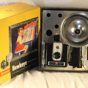 May include: A yellow box with a picture of a girl painting on an easel. The box says "Brownie Hawkeye Flash Outfit No. 1272". The box is open and shows a black and silver camera with a flash attachment.