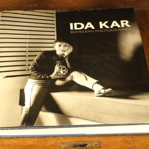 May include: A monochrome book cover with the title "IDA KAR Bohemian Photographer". The image shows a person with a camera. The book is on a wooden surface.