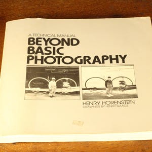 May include: A technical manual titled "Beyond Basic Photography" with black text on a cream-colored cover. The cover features two black and white images of people with bicycles. The author is Henry Horenstein.