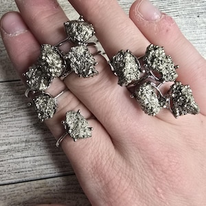 May include: Silver-toned rings featuring rough, textured pyrite stones. The rings have a simple band design with the pyrite stones securely mounted. The pyrite stones have a metallic, speckled appearance, with a mix of silver and gold tones.
