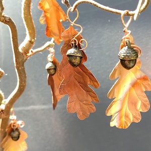 May include: A set of hanging ornaments featuring metallic copper-coloured oak leaves paired with small, textured bronze-toned acorn caps. Each piece is suspended by a coiled copper wire loop, showcasing detailed leaf veins and a natural autumn colour palette.