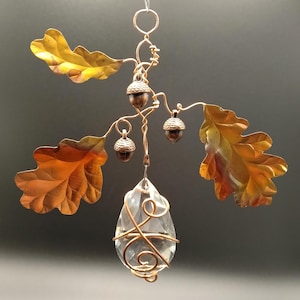 May include: A hanging ornament featuring a clear, teardrop-shaped crystal wrapped in copper wire. The crystal is suspended from a copper wire structure decorated with three metallic oak leaves in shades of gold and bronze, and two small acorns.