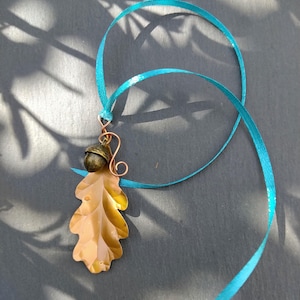 May include: A pendant featuring a light brown, translucent oak leaf shape attached to a small, dark metallic acorn charm. The charm is connected by a copper-coloured wire spiral to a bright turquoise ribbon that loops through the top of the wire.