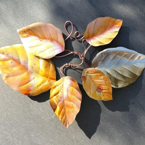 May include: A decorative wall hanging featuring five metal leaves in shades of yellow, orange, and copper. The leaves are connected by thin, twisted copper wire stems that form a central cluster, creating a naturalistic arrangement of foliage.