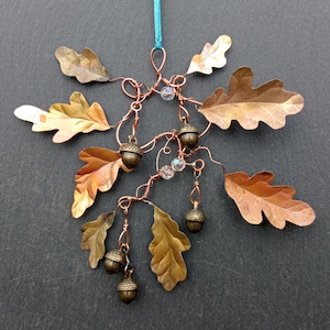 May include: A handmade copper ornament with oak leaves and acorns. The leaves have a bronze and gold finish, and the acorns are a dark bronze colour. Clear beads are interspersed, and a teal string is used for hanging.