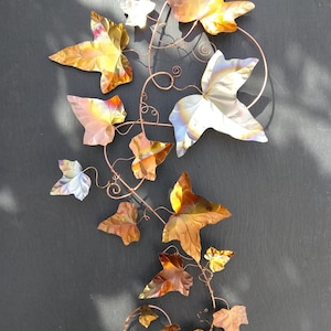 May include: A wall-mounted metal sculpture featuring a vine with multiple leaves in shades of copper, bronze, and iridescent silver. The leaves are attached to thin, curved copper wire stems that form a trailing, organic pattern with small curls.