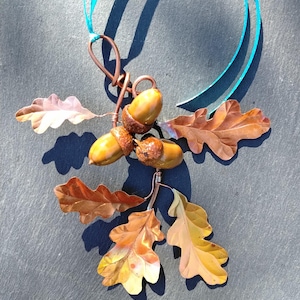 May include: A decorative hanging ornament featuring three realistic brown acorns with textured caps attached to a copper wire frame. The ornament includes five metallic copper-coloured oak leaves and a bright turquoise ribbon for hanging purposes.