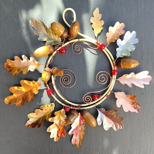 May include: A circular wall hanging featuring two concentric light-coloured rings adorned with metallic oak leaves in shades of copper, bronze, and silver. The design includes several brown acorns and small red beads attached with copper wire spirals.