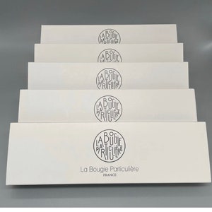 Peut inclure: Une pile de six boîtes blanches avec le texte "La Bougie Particulière" et "France" imprimé sur la boîte du bas. Chaque boîte a un logo circulaire avec le texte "La Bougie Particulière" dans une police de caractères stylisée.