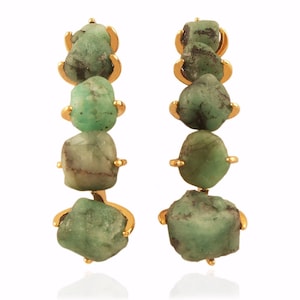 May include: A pair of gold earrings, each featuring five emerald green stones. The stones are irregularly shaped and have a rough, natural texture.