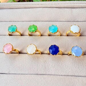 May include: Eight gold-toned rings arranged in two rows, each featuring a single round, faceted gemstone. The stones display a variety of colors including mint green, bright green, turquoise, white, soft pink, cream, deep blue, and light blue.