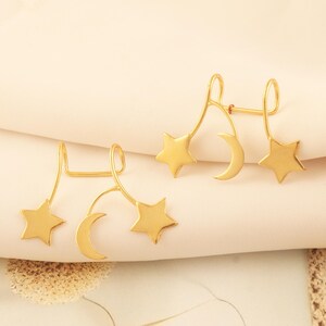 May include: Gold-colored celestial earrings featuring a star and crescent moon design. The earrings have a curved bar with a star at the top and a crescent moon below. The earrings are displayed on a cream-colored surface.