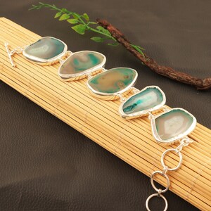 Puede incluir: Un brazalete de plata con cinco piedras de ágata verde y blanca. Las piedras son de forma ovalada y tienen un aspecto natural y orgánico. El brazalete tiene un cierre de mosquetón.