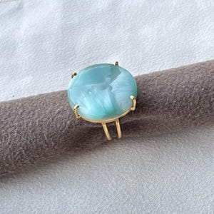May include: A gold ring featuring a large, round, polished light blue stone with white marbled patterns. The stone is secured by four gold prongs, and the band consists of two thin, parallel gold wires that meet at the base of the setting.