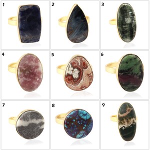 Pode incluir: Nove anéis de ouro com diferentes engastes de pedras preciosas. Os anéis estão dispostos em três filas de três. As pedras preciosas são todas únicas e têm padrões e cores diferentes.