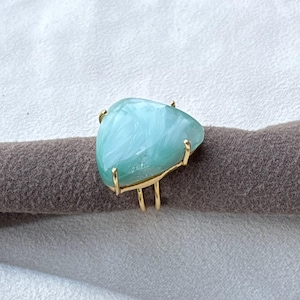 May include: A gold-toned ring featuring a large, triangular, light blue marbled stone. The stone is held in place by four thin gold-toned prongs, and the band is composed of two parallel gold-toned wires that meet at the base of the setting.