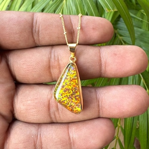 May include: A gold-toned necklace featuring a triangular pendant with a vibrant, multi-colored iridescent inlay. The pendant is suspended from a delicate gold-toned chain, showcasing a textured surface with flecks of orange, yellow, and red.