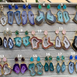 Puede incluir: Una colección de pendientes de piedras preciosas con ribete dorado. Los pendientes presentan varios colores de ágata, incluyendo azul, verde, morado y marrón. Cada par tiene una piedra preciosa diferente en la parte superior, como una piedra azul en forma de lágrima.