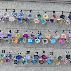 May include: A display of 34 gemstone rings with silver bands. The rings showcase a variety of colours and patterns, including blue, purple, orange, and green. Each ring is numbered for easy selection.