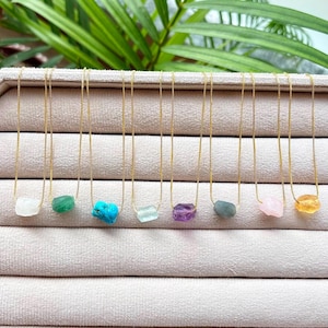 May include: Eight delicate gold-tone box chains, each featuring a single raw, uncut gemstone pendant. The stones display a variety of colors including white, green, turquoise, light blue, purple, grey, pale pink, and yellow in a horizontal row.