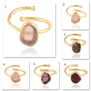 May include: A collection of six gold-toned open-ended rings, each featuring a single raw, irregularly shaped gemstone in various colors including pale pink, dark brown, light peach, purple, and deep red, set in a simple metal band.