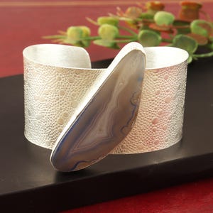 May include: A silver cuff bracelet with a textured surface and a blue agate stone accent.