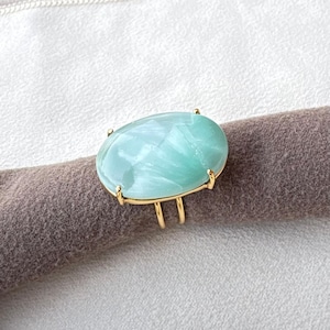 May include: A ring featuring a large, oval-shaped, light mint green stone with a smooth, polished surface. The stone is set in a gold-toned metal prong setting with a split band design that wraps around the finger.