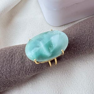 May include: A gold-toned ring featuring a large, oval-shaped, smooth, light green stone with subtle white marbled veining. The stone is secured by four small prongs on a simple, thin, metallic band that rests horizontally.