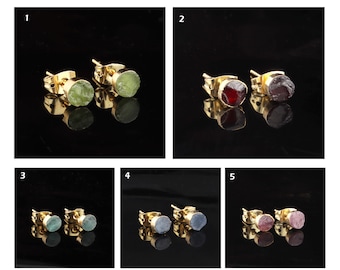 Puede incluir: Cinco pares de pendientes de botón de oro con piedras preciosas de diferentes colores. Los pendientes se muestran sobre un fondo negro. Las piedras preciosas son verdes, rojas, azules y rosas.