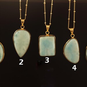 May include: Five gold-coloured necklaces with various pendant shapes. The pendants are pale blue and white, with gold-coloured frames. The necklaces are displayed against a black backdrop. Each pendant is numbered from 1 to 5.