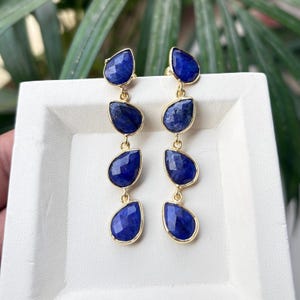 Puede incluir: Un par de pendientes dorados con tres piedras preciosas azules oscuras facetadas en forma de lágrima, suspendidas de una piedra más pequeña y similar. Los pendientes están sobre un fondo blanco con follaje verde.