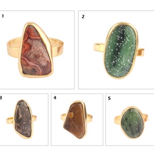 May include: Five gold-toned rings with unique stone settings. The stones vary in color, including red, green, brown, and black. Each ring features a different shaped stone, showcasing natural patterns and textures. The rings are displayed on a white background.