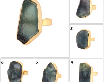 Könnte beinhalten: Ein goldfarbener Ring mit einem großen, unregelmäßig geformten, teal-farbenen Edelstein. Der Edelstein ist in einer goldenen Lünette gefasst, was einen auffälligen Kontrast erzeugt. Das Ringband ist ebenfalls goldfarben. Das Design ist kühn und auffällig.