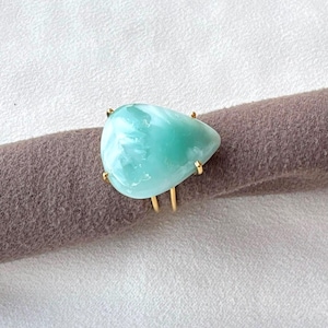 May include: A gold ring featuring a large, smooth, teardrop-shaped light blue stone. The gemstone has a polished surface with subtle white inclusions and is secured by four small gold prongs on a thin, delicate gold band.