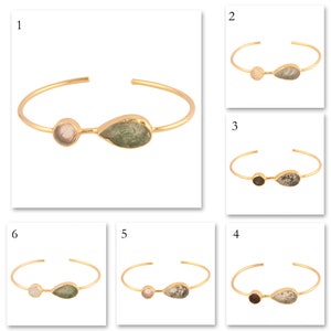 Könnte beinhalten: Goldfarbenes Armreif-Armband mit zwei Edelstein-Akzenten. Einer ist rund und der andere tropfenförmig. Die Edelsteine sind hellrosa und grün. Das Armband ist unten offen.