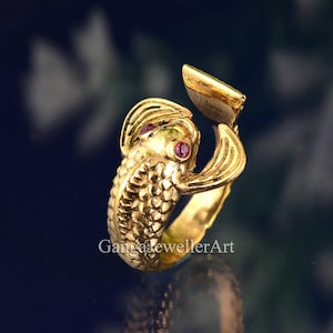 May include: Gold ring shaped like a fish with ruby red eyes. The fish is curled around the finger and the tail is pointing up.
