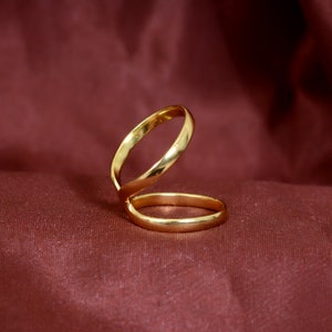 May include: A gold ring with a unique design, featuring two interlocking bands. The ring is lying on a burgundy fabric background.
