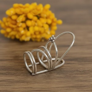 May include: A silver-colored ring holder with a cluster of small beads at the top. The holder has a series of curved, parallel bands. A blurred cluster of yellow flowers is in the background. The ring holder is on a wooden surface.