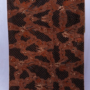 May include: A brown and black batik fabric with a floral pattern. The fabric features a repeating design of stylized flowers and leaves, with a dark background and lighter brown accents. The fabric is likely used for clothing or home decor.
