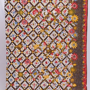 Könnte beinhalten: Rechteckiger Stoff mit einem sich wiederholenden Muster aus braunen Rauten, die jeweils ein Blumendesign in Rot, Gelb, Blau und Weiß enthalten. Ein Rand aus Blumen und Blättern in ähnlichen Farben umrahmt den Rand. Der Stoff hat einen roten und gelben Rand.