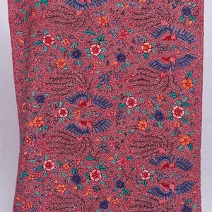 Op de afbeelding: Een rode stof met een gedetailleerd bloemen- en vogelpatroon. Het ontwerp toont gestileerde vogels met blauwe vleugels en rode accenten, afgewisseld met rode, oranje en blauwe bloemen. De achtergrond is een gespikkeld patroon.