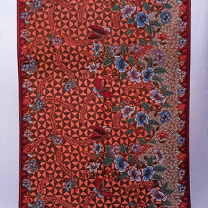 May include: A rectangular textile with a vibrant floral pattern. The design features a geometric background in shades of orange and brown, with detailed floral and butterfly motifs in blue, red, and purple. The edges are bordered with a deep red.