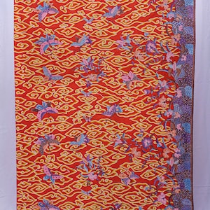 Op de afbeelding: Een levendig rode stof met een herhalend patroon van gestileerde vlinders, vogels en wolkvormige figuren in geel, roze en blauw. Een bloemenrand in paarse en blauwe tinten omlijst het ontwerp. Geschikt voor kleding of woondecoratie.