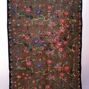 May include: A rectangular textile with a dark brown background, featuring a repeating pattern of small, light brown circles. The fabric showcases a vibrant floral design with red, blue, and green flowers and birds. The edges are framed with a black border.