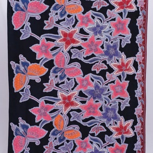 以下が含まれることがあります： 黒色のテキスタイルで、花柄模様です。ピンク、赤、オレンジ、紫の花と蝶のような形がデザインされています。縁は濃い赤色のボーダーでトリミングされています。